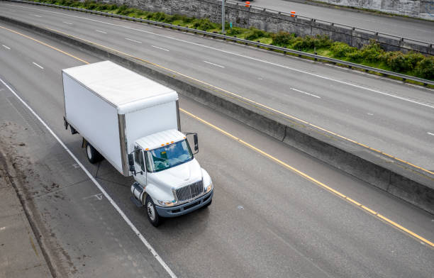 Popular size big rig day cab white semi truck with long box trailer making local commercial cargo delivery at urban city driving on the highway road with exit intersection
